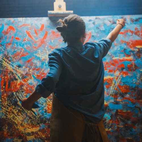 Talented Female Artist Works on Abstract Oil Painting, Using Paint Brush She Creates Modern Masterpiece. Dark and Messy Creative Studio where Large Canvas Stands on Easel Illuminated.